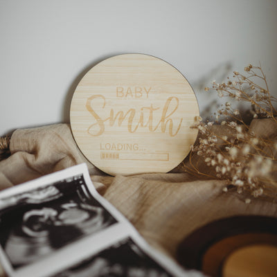 A light bamboo round plaque saying Baby Smith Loading..... with a loading icon underneath. Sitting next to an ultrasound photo.