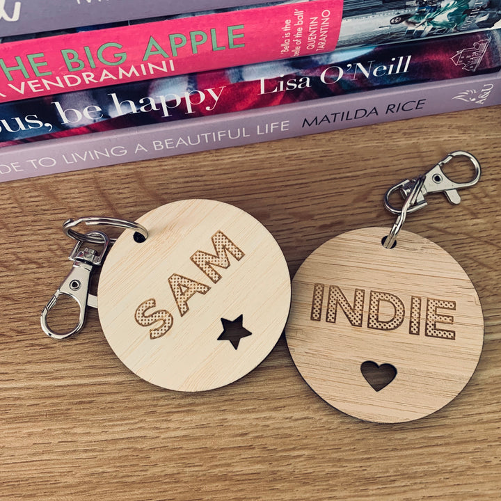 Two round wooden Polka Dot Key Rings rest on a table: one is engraved with SAM and a star, the other with INDIE in a polka dot font and a heart. Stacked books with colorful spines and visible titles sit behind them.