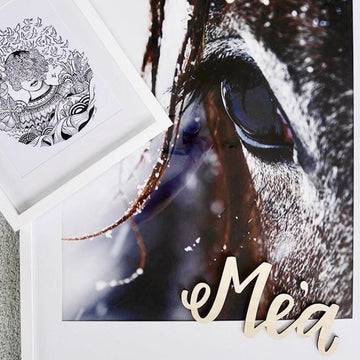 A close-up of a horse’s eye with the Lovely Lettering Handlettered Wall Name spelling “Mea” in cursive wooden letters. A small, black-and-white abstract drawing with a woman’s face overlaps one corner of the photo.