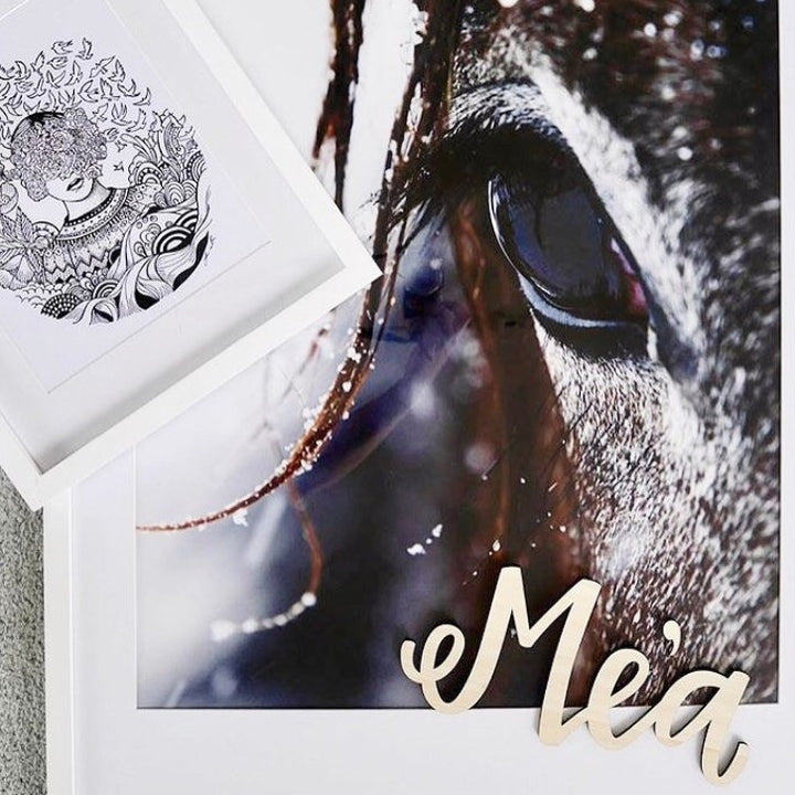 A close-up of a horse’s eye with the Lovely Lettering Handlettered Wall Name spelling “Mea” in cursive wooden letters. A small, black-and-white abstract drawing with a woman’s face overlaps one corner of the photo.