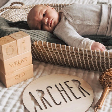 A baby in a woven basket wears a light outfit on a bed, with wooden blocks showing "01 weeks old" and a Personalised Natural Names Plaque featuring "Archer," adding unique decor to the cozy setting.