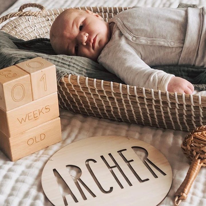 A baby in a woven basket wears a light outfit on a bed, with wooden blocks showing "01 weeks old" and a Personalised Natural Names Plaque featuring "Archer," adding unique decor to the cozy setting.
