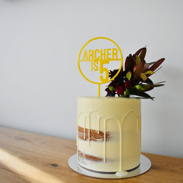 A semi-naked cake with white icing drips, topped with a cluster of dark red and green flowers and a yellow Target Personalised Cake Topper (other colours available), sits on a wooden table against a plain white wall.