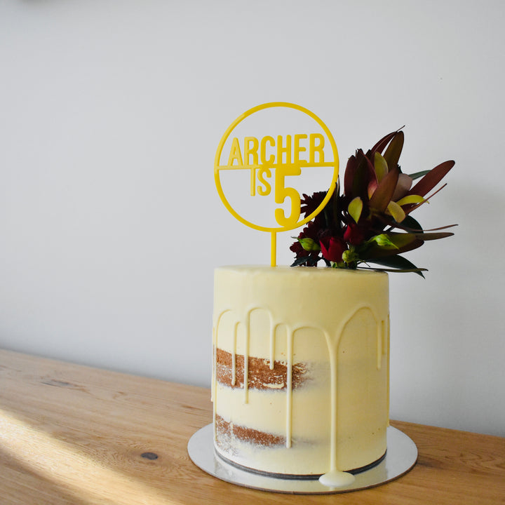 A semi-naked cake with white icing drips, topped with a cluster of dark red and green flowers and a yellow Target Personalised Cake Topper (other colours available), sits on a wooden table against a plain white wall.