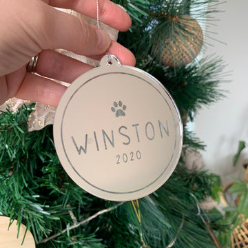 A hand holds an Etched Pets Christmas Bauble, a round, reflective ornament with a paw print and personalised text, displayed in front of a decorated green Christmas tree.