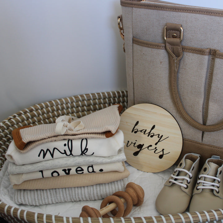 A woven basket holds folded neutral baby clothes, a Personalised Announcement Plaque reading “baby vigers,” baby milestone cards, a pair of baby shoes, wooden pacifier holders, and a beige diaper bag with pockets.