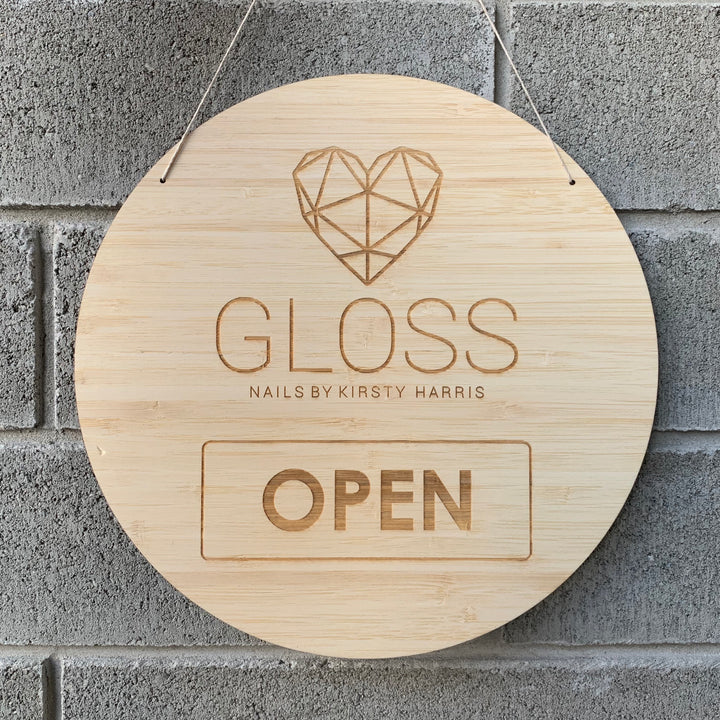 A light bamboo round sign saying open, with Gloss Nails logo engraved. Hanging against a grey brick wall.