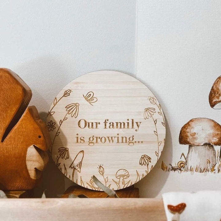 A round bamboo saying Our family is growing... with a floral border by Chloe Mytton, sitting on a shelf.