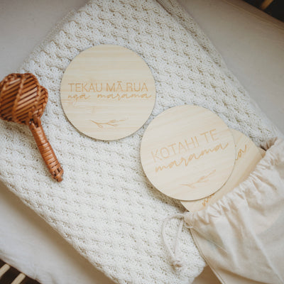 A round light bamboo set of baby milestones in a white calico bag, lying on a baby blanket with a rattle. The two visible signs say Tekau mā rua ngā marama (twelve months) and kotahi te marama (one month).