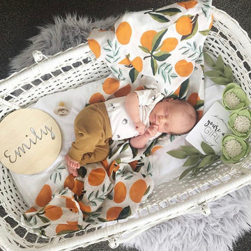 A newborn rests in a white bassinet with an orange-patterned blanket, beside a Personalised Natural Names Plaque reading "Emily," a "you&