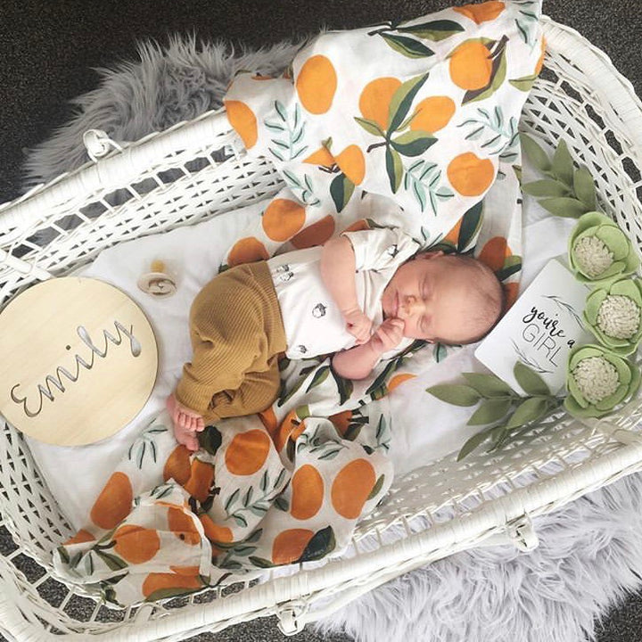 A newborn rests in a white bassinet with an orange-patterned blanket, beside a Personalised Natural Names Plaque reading "Emily," a "you&