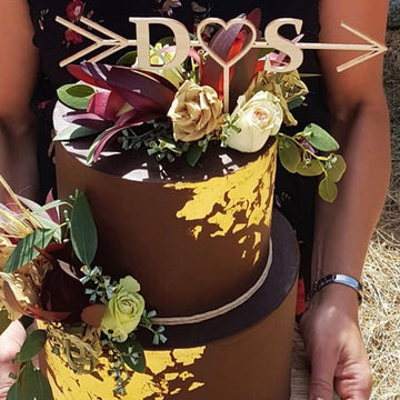 An arrow shaped cake topper in light bamboo, with a heart and the letters D and S. Sitting on a brown cake with gold embellishments, and a floral arrangement.
