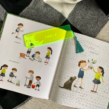 A neon green clear acrylic bookmark with florals and the name Indie etched on, with a green tassel, sitting atop a children&