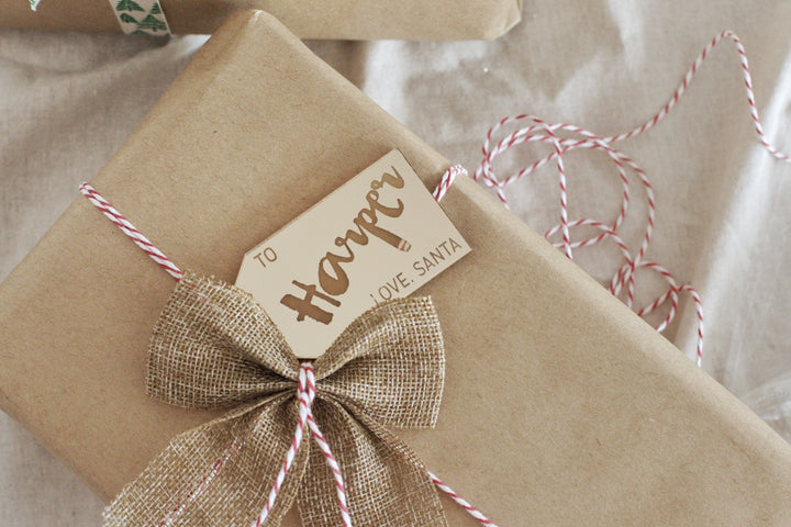 A gift wrapped in brown paper with red and white twine, finished with a burlap bow and our Personalised Gift Tags reading “To Harper, Love, Santa.”.