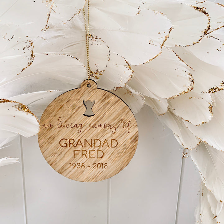 The In Loving Memory Christmas Bauble is a round wooden ornament engraved with "In loving memory of GRANDAD FRED 1938 - 2018", featuring an angel cutout, hung by a gold string before white feathers with gold tips.