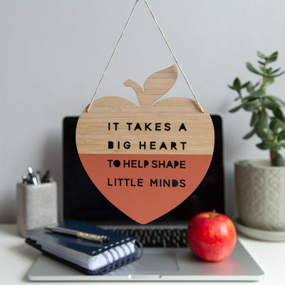 A heart shaped apple in bamboo, half painted red saying "It takes a big heart to help shape little minds". Hanging in front of a teachers desk with laptop, stationary and an apple