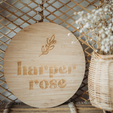 The Personalised La Petite Stem Plaque, a round bamboo sign with an engraved branch and custom text, rests against a woven lattice near a wicker basket of dried white flowers—a charming birth announcement or personalised display.