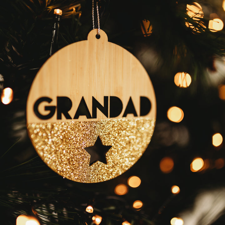The Glittered Christmas Bauble is a round bamboo ornament featuring GRANDAD in bold letters, a glittery gold bottom half with a star cutout, and hangs beautifully on the tree amid twinkling lights.