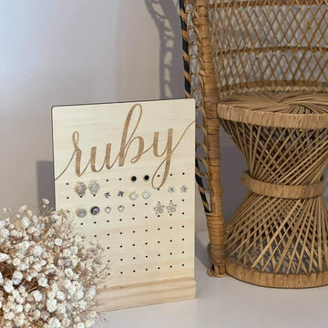 The Personalised Earring Holder, engraved with the name Ruby, displays several pairs of earrings. It sits next to a wicker chair and a bouquet of small white flowers on a light-colored surface.