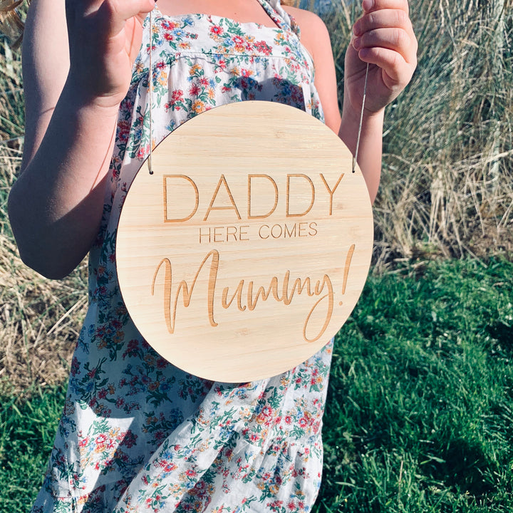A girl holding a round light bamboo sign saying Daddy here comes Mummy!
