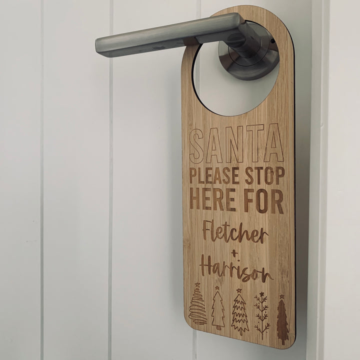 The Santa Stop Here Door Hanger, personalised with “SANTA PLEASE STOP HERE FOR Fletcher + Harrison” and engraved tree images, adds a charming touch to your Christmas tradition when hung on a silver handle against any door.