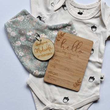 A dark bamboo rectangle birth announcement card saying Hello my name is. Born, Time, Weight and Length. Laying next to a baby outfit, and light bamboo amazing midwife at your cervix keyring.