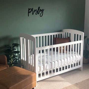 A white baby crib with a brown pillow and blanket sits against a green wall, displaying a Black Acrylic Wall Name (other sizes and fonts available) in Luxe font. Green potted plants and natural sunlight complete the cozy room.
