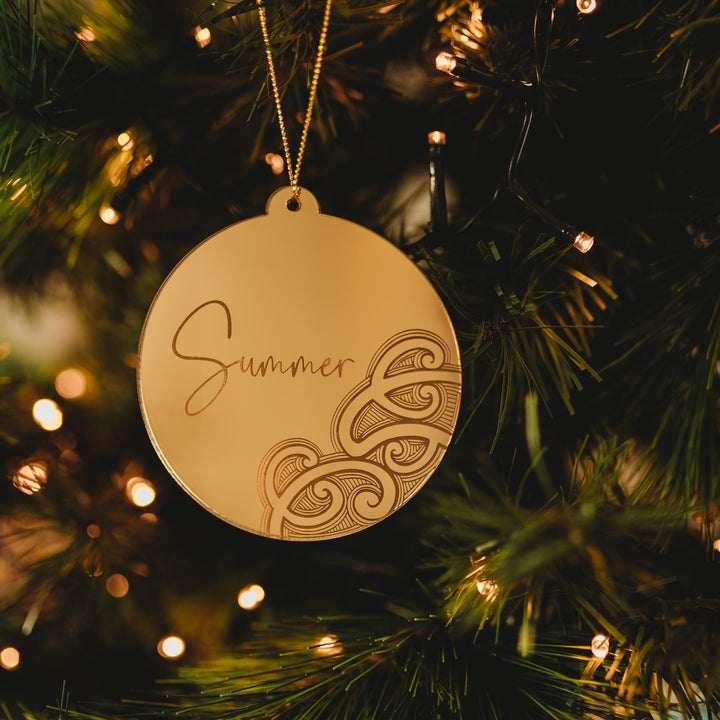 A gold round Aotearoa Kirihimete Bauble engraved with "Summer" and a decorative pattern hangs on a Christmas tree among green branches and warm white lights—a stunning piece by Ani Huia Art.