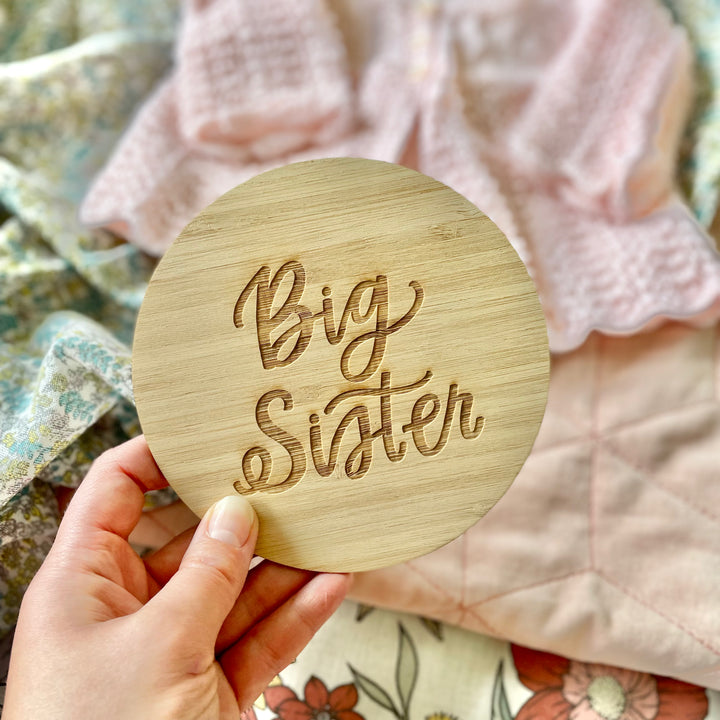 A hand holds the Lovely Lettering Sibling Announcement Card engraved with "Big Sister"—ideal for announcing a big sister. Pastel quilts and a pink knitted sweater in the background enhance this charming personalised decor scene.
