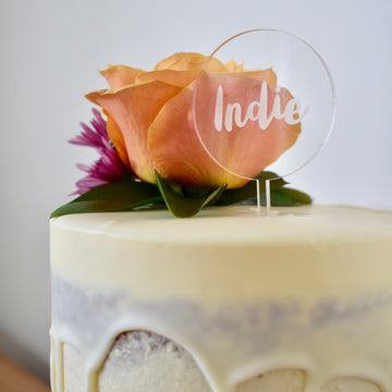 A round clear acrylic cake topper saying Indie. Sitting on a white drip cake with floral arrangement. 