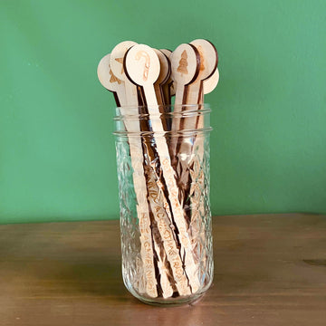The Activity Advent Calendar Set includes a glass jar filled with wooden drink stirrers, each engraved with designs and text, ideal for festive family countdown activities, displayed on a wooden surface against a green wall.