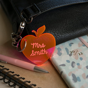 A Teacher Key Ring, apple-shaped and engraved with "Mrs Smith," hangs on a black bag. Nearby sit a pink pen, spiral notebook, and pastel 2022 planner—a thoughtful personalized teacher gift.