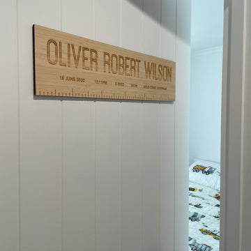 A Personalised Birth Ruler featuring OLIVER ROBERT WILSON and birth details hangs on a white wall by an open door, revealing a bedroom with a bed dressed in a vehicle-themed duvet.