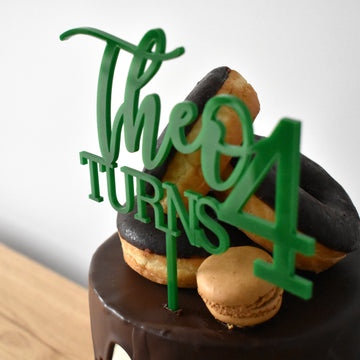 Green acrylic cake topper saying Theo Turns 4, sitting on a chocolate drip cake with donuts.