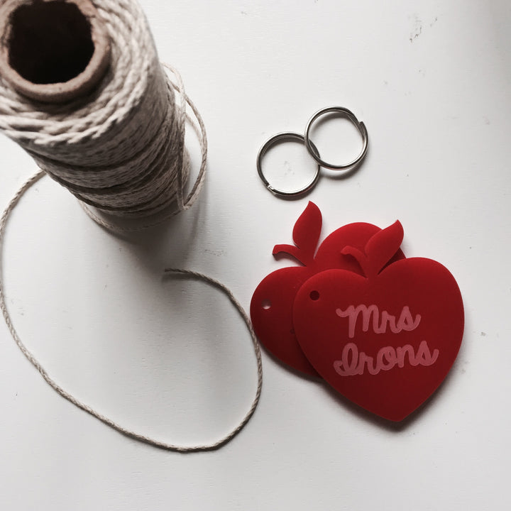 The Teacher Key Ring includes a spool of twine, two metal key rings, and two red apple-shaped tags—one labelled "Mrs Irons"—displayed on a white surface, ideal for personalized teacher keychains or thoughtful gifts.