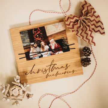 The Christmas Photo Holder, made in New Zealand, displays a family photo with “Christmas Memories,” accented by a red and beige bow, pinecones, white ornaments, and red-white string on a light background.