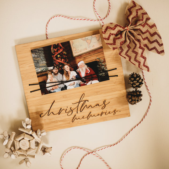 The Christmas Photo Holder, made in New Zealand, displays a family photo with “Christmas Memories,” accented by a red and beige bow, pinecones, white ornaments, and red-white string on a light background.