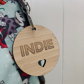 The Polka Dot Key Ring features "INDIE" in bold polka dots and a heart cutout on wood, hanging from a metal ring attached to a cartoon-themed fabric bag. Ideal for anyone who loves quirky, personalised accessories.