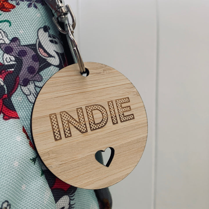 The Polka Dot Key Ring features "INDIE" in bold polka dots and a heart cutout on wood, hanging from a metal ring attached to a cartoon-themed fabric bag. Ideal for anyone who loves quirky, personalised accessories.