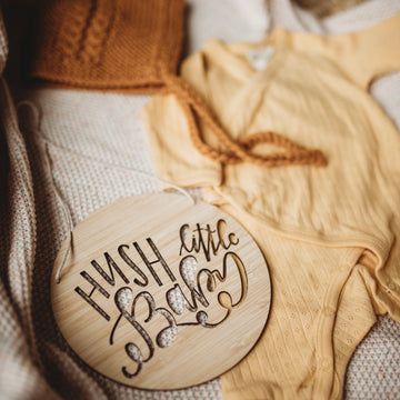 A round light bamboo plaque saying hush little baby, sitting with a yellow baby outfit and white blanket.