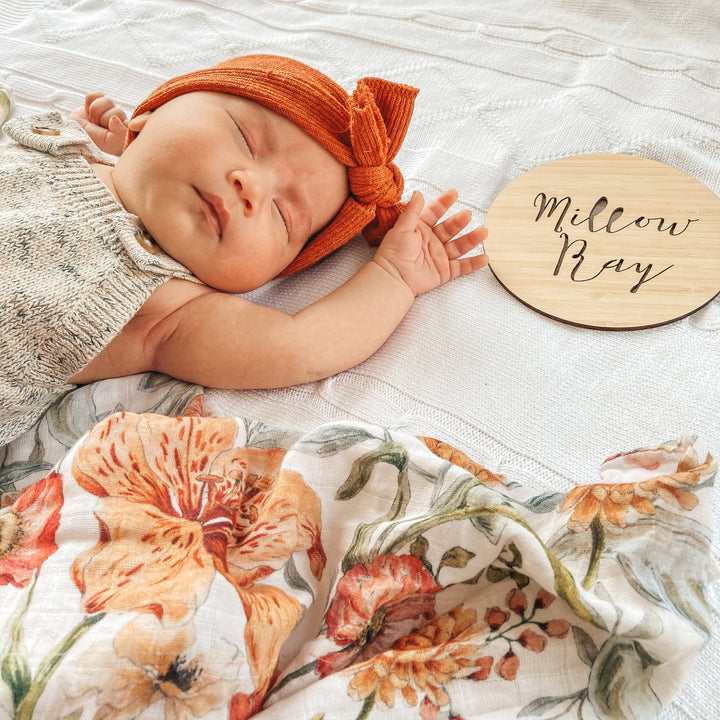 A baby wearing an orange headband lies on a white textured blanket beside a Personalised Announcement Plaque that reads “Willow Bay.” An orange floral blanket is partially visible.