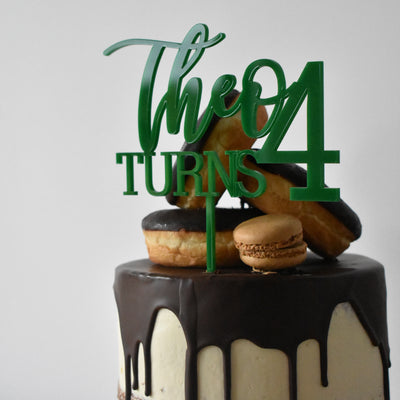 Green acrylic cake topper saying Theo Turns 4, sitting on a chocolate drip cake with donuts.