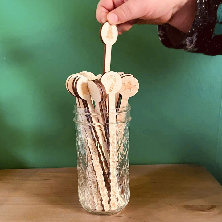A hand holds a wooden stir stick with a tree design above a jar of the Activity Advent Calendar Set—engraved family activity sticks featuring unique symbols—perfect for a Christmas DIY project against a green wall background.