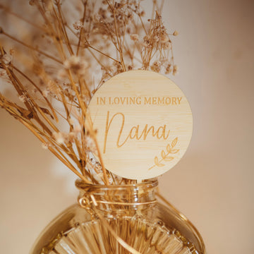 A round light bamboo planter saying In Loving Memory Nana, with a laurel engraved. Sitting with a dried floral arrangement. 