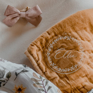 A Personalised Frosted Christmas Bauble featuring the name Ellie inside a white floral wreath, displayed beside a pink bow, a floral-patterned cloth, and keepsake gift packaging on a light surface.