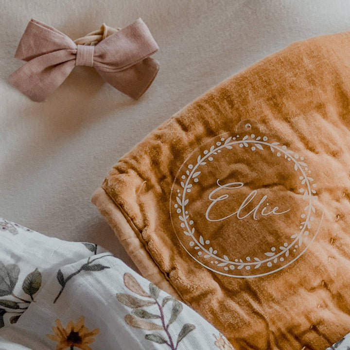 A Personalised Frosted Christmas Bauble featuring the name Ellie inside a white floral wreath, displayed beside a pink bow, a floral-patterned cloth, and keepsake gift packaging on a light surface.