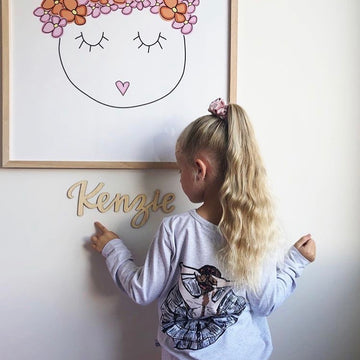 A young girl with long blond hair in a ponytail points to the Lovely Lettering Handlettered Wall Name reading "Kenzie" on the wall, standing beneath a simple framed drawing of a face with closed eyes and a flower crown.