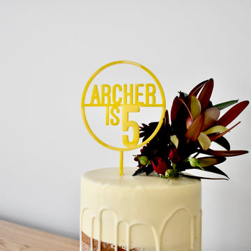 A white frosted cake features Target Personalised Cake Topper (other colours available) reading ARCHER IS 5 in yellow acrylic, and is decorated with red and green flowers on the right side, set against a plain white background.
