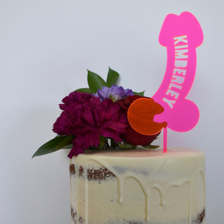 A cake with white icing and flower decorations, topped with a bright pink BDE Personalised Cake Topper (other colours available) spelling KIMBERLEY—perfect for adding fun and a personalised touch to your hen’s night celebration.