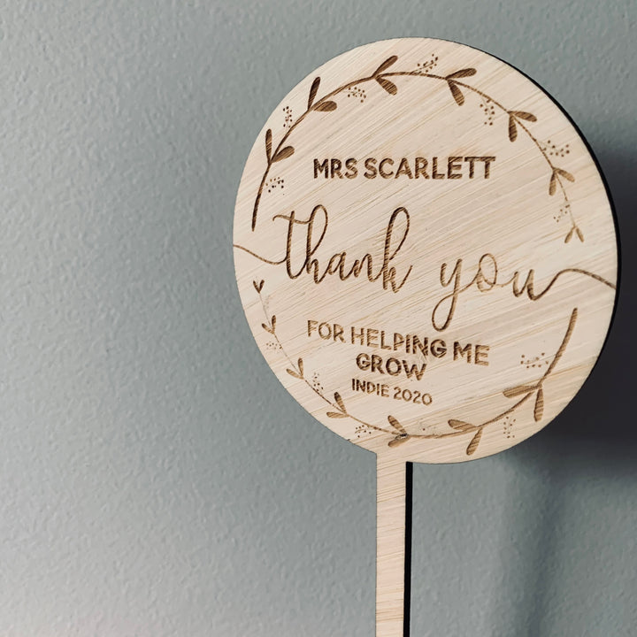 The Personalised Wreath Teacher Planters feature a round wooden plaque on a stick, engraved with “Mrs Scarlett thank you for helping me grow Indie 2020” and a leafy border—an ideal teacher gift displayed against a light gray wall.